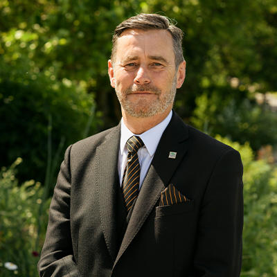A male funeral director wears a dark jacked and black and gold suit and smiles gently at the camera